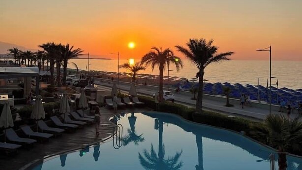 A resort pool area with lounge chairs and umbrellas, a sunset view, and a beach in the background. - Aegean Pearl (Rethymno)
