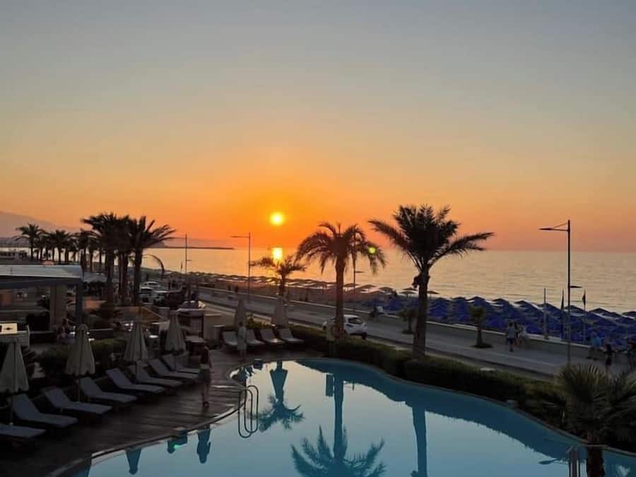 Een zwembadgedeelte van een resort met ligbedden en parasols, uitzicht op een zonsondergang en een strand op de achtergrond.