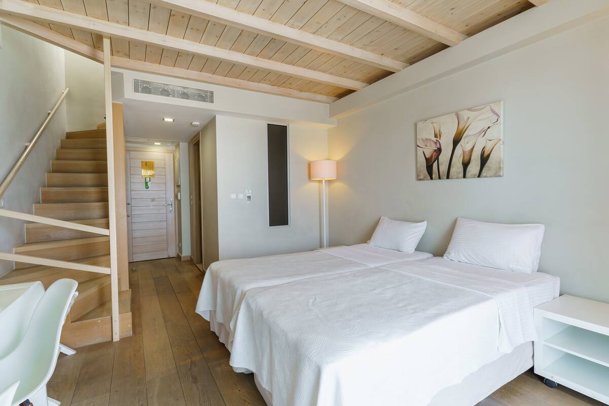 A bedroom with two beds, a wooden staircase, a white wall with a floral painting, and a lamp.