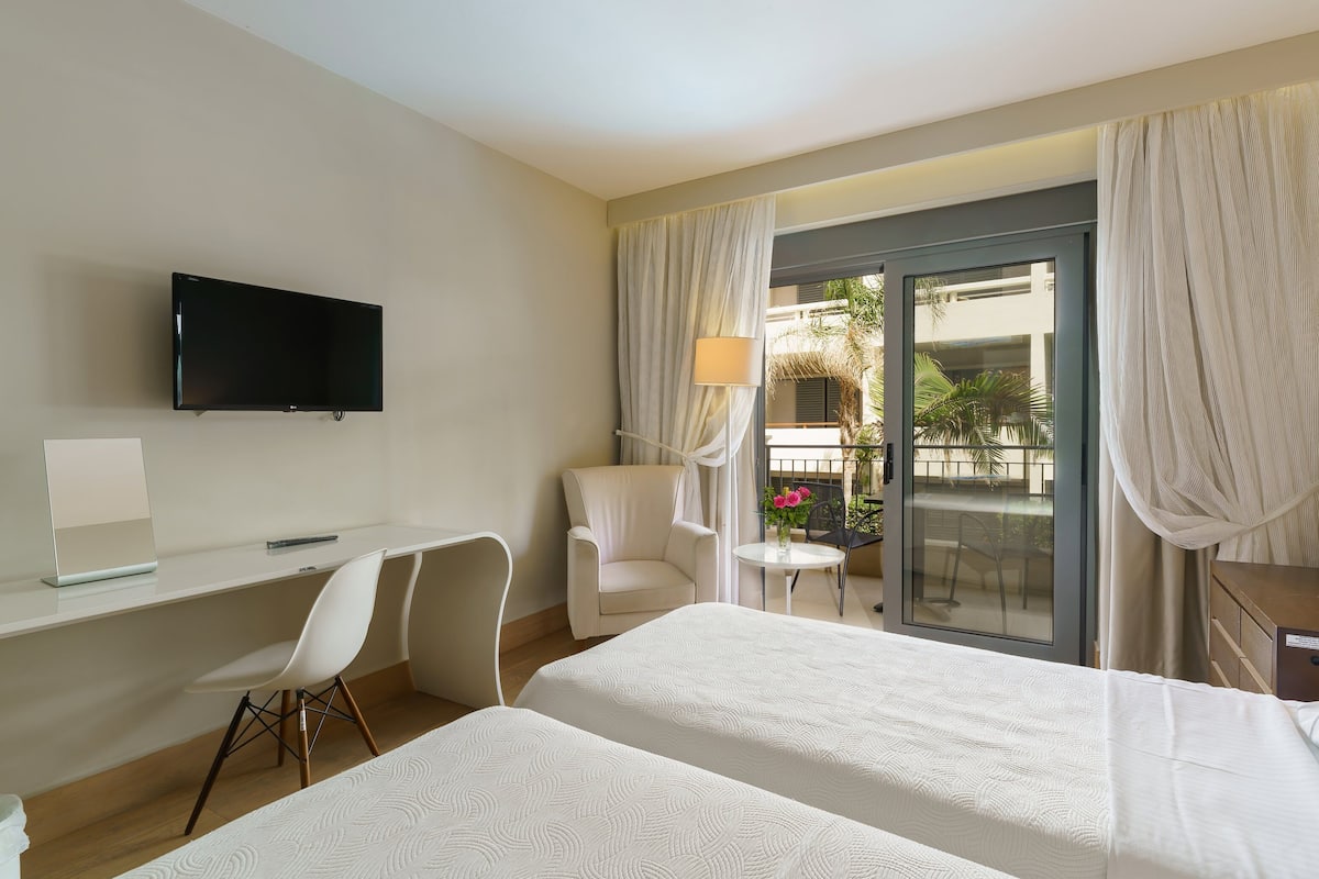 A hotel room with two beds, a desk with a chair, a TV mounted on the wall, and a balcony with curtains.