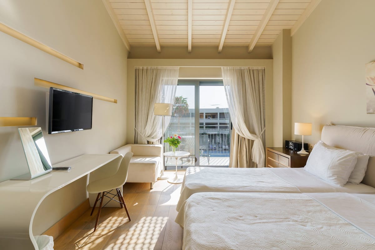 A hotel room with a desk, two beds, a TV, and a balcony with a table and chairs.