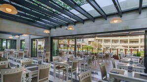 A spacious outdoor dining area with multiple tables and chairs, a covered section with a ceiling structure, and large windows providing views of the surrounding area. - Aegean Pearl (Rethymno)