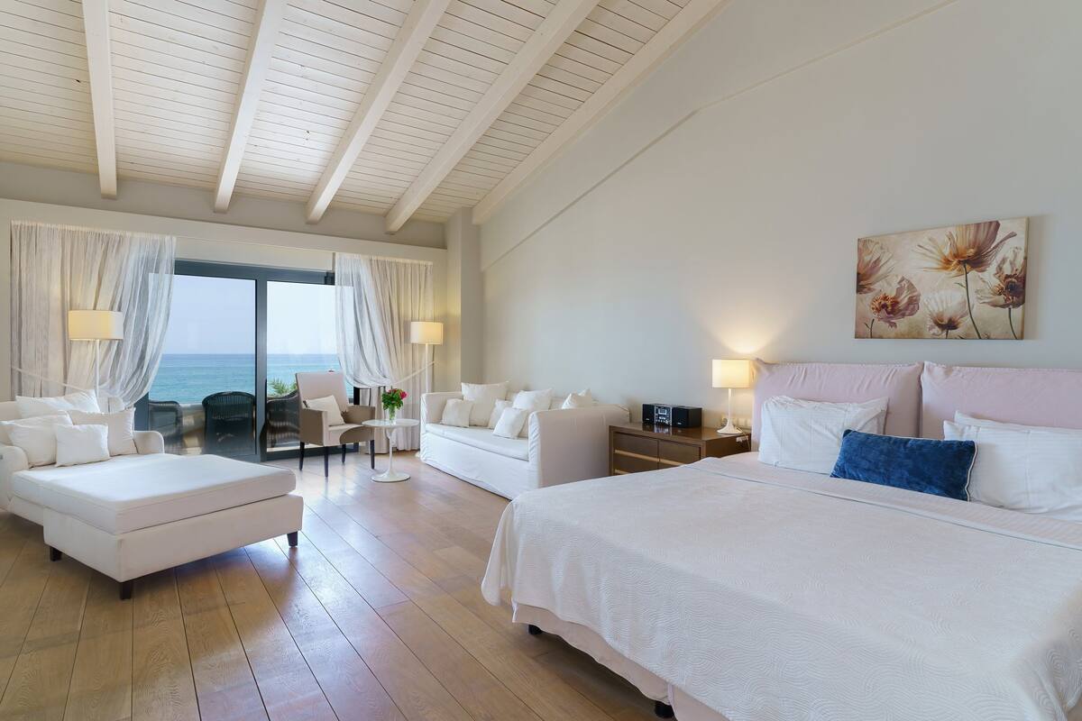 A hotel room with a large bed, a sofa, a chair, and a table. There is a view of the sea through the windows.