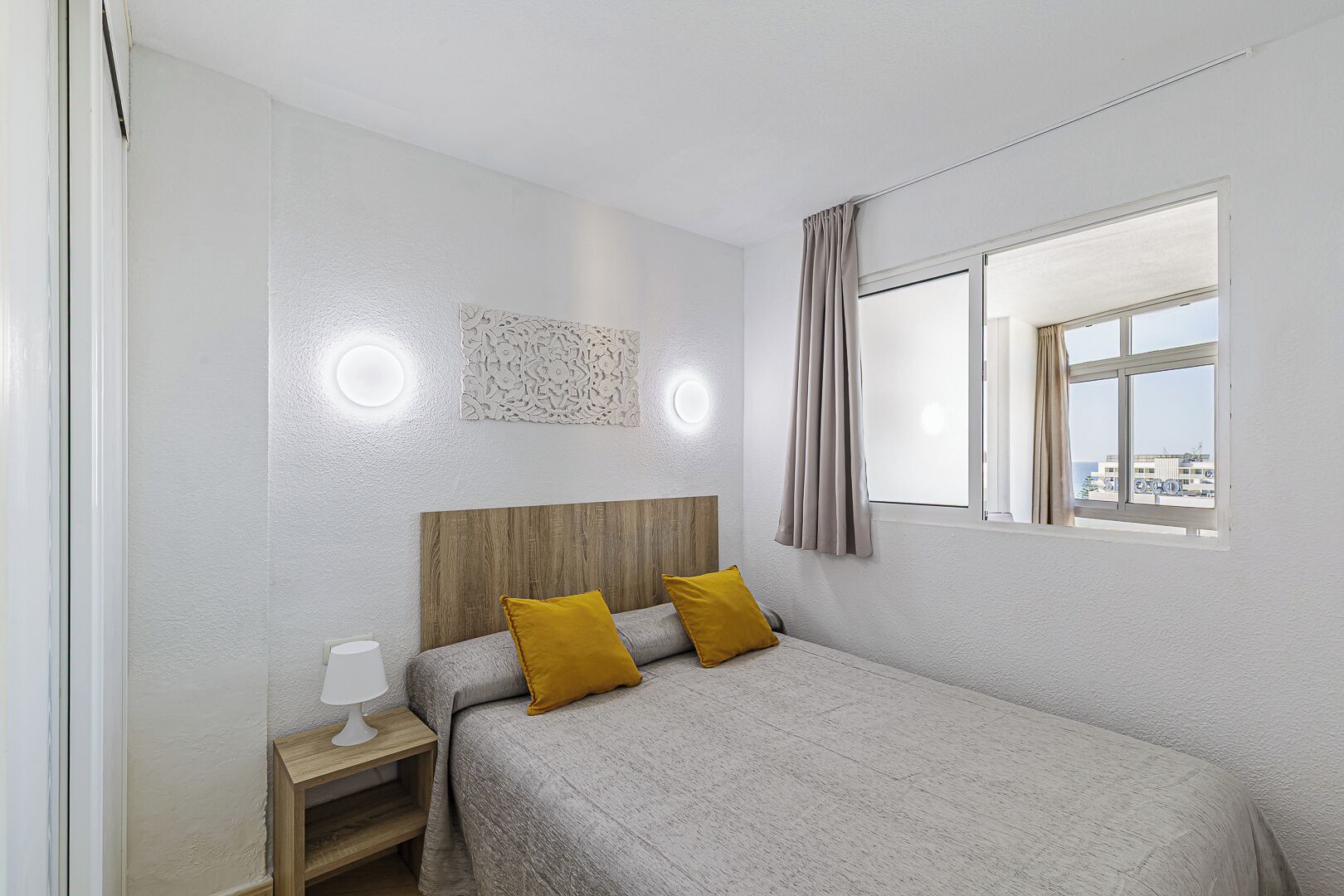 A bedroom with a bed, a nightstand, a wall-mounted light, a window with curtains, and a decorative wall piece.