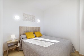 Ein Schlafzimmer mit Holz-Kopfstütze, einem Bett mit grau-weißem Muster, zwei senfgelben Kissen, einem Nachttisch mit Lampe und zwei Wandleuchten.