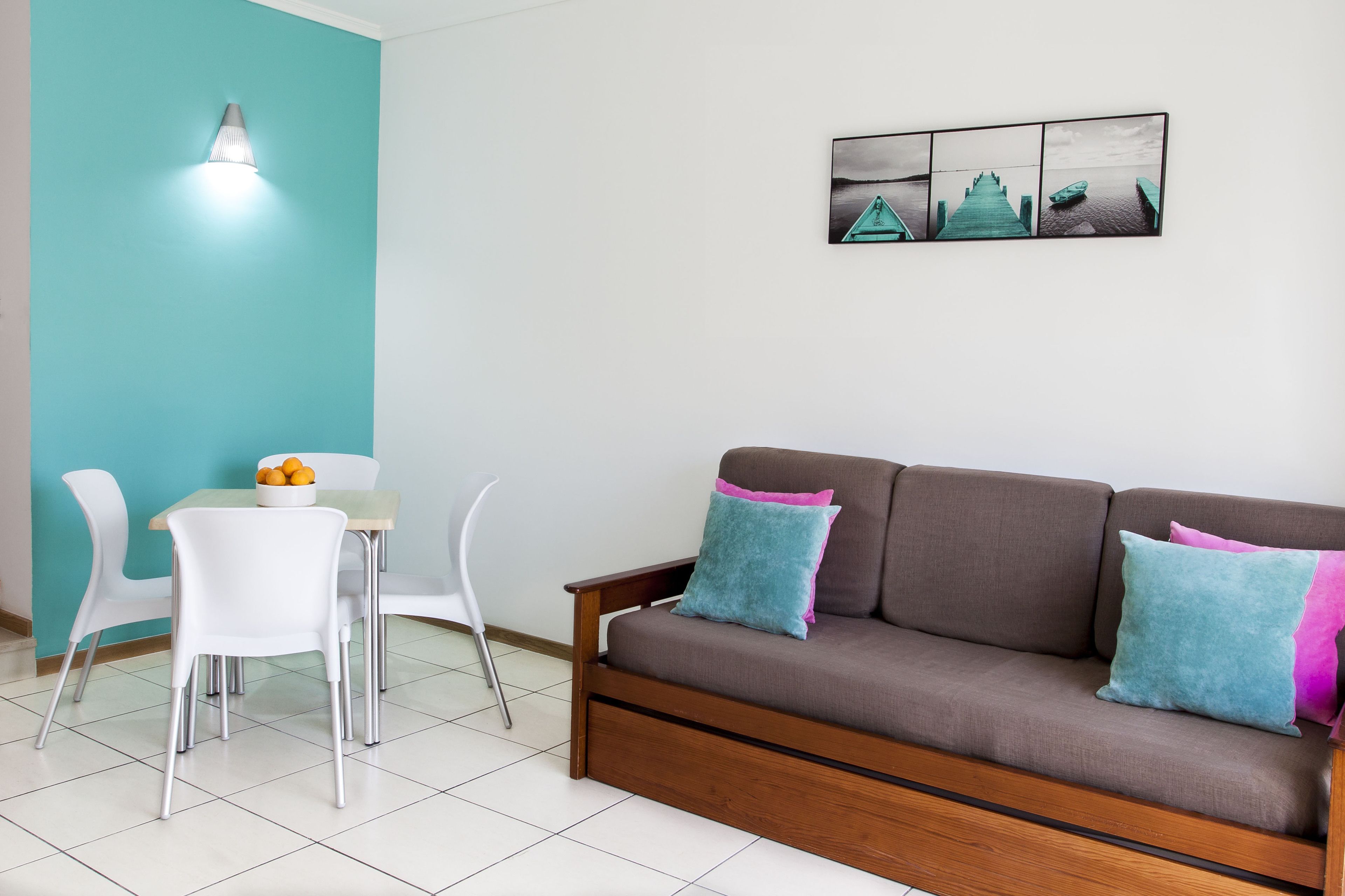 Uma sala de estar com um sofá marrom, cadeiras brancas, uma mesa pequena com uma tigela de frutas, uma luminária fixada na parede e uma imagem de um cais.