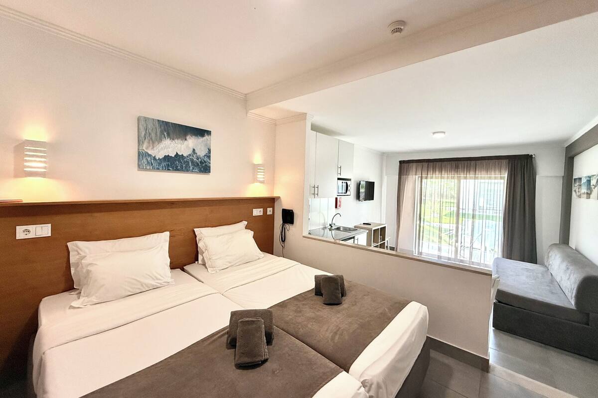 A hotel room with a double bed, a small sofa, a bathroom with a sink and mirror, and a window with curtains.