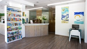 A reception area with a wooden reception desk, a bookshelf filled with books, and a computer on a small table. - BaySide Salgados (Albufeira)