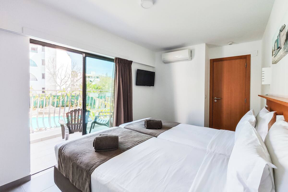 A hotel room with a bed, a TV, a door, and a balcony with a pool.