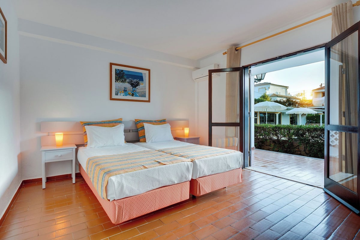 Camera da letto con due letti, un comodino con lampada e un'ampia porta-finestra scorrevole che si apre su una zona esterna con ombrelloni.