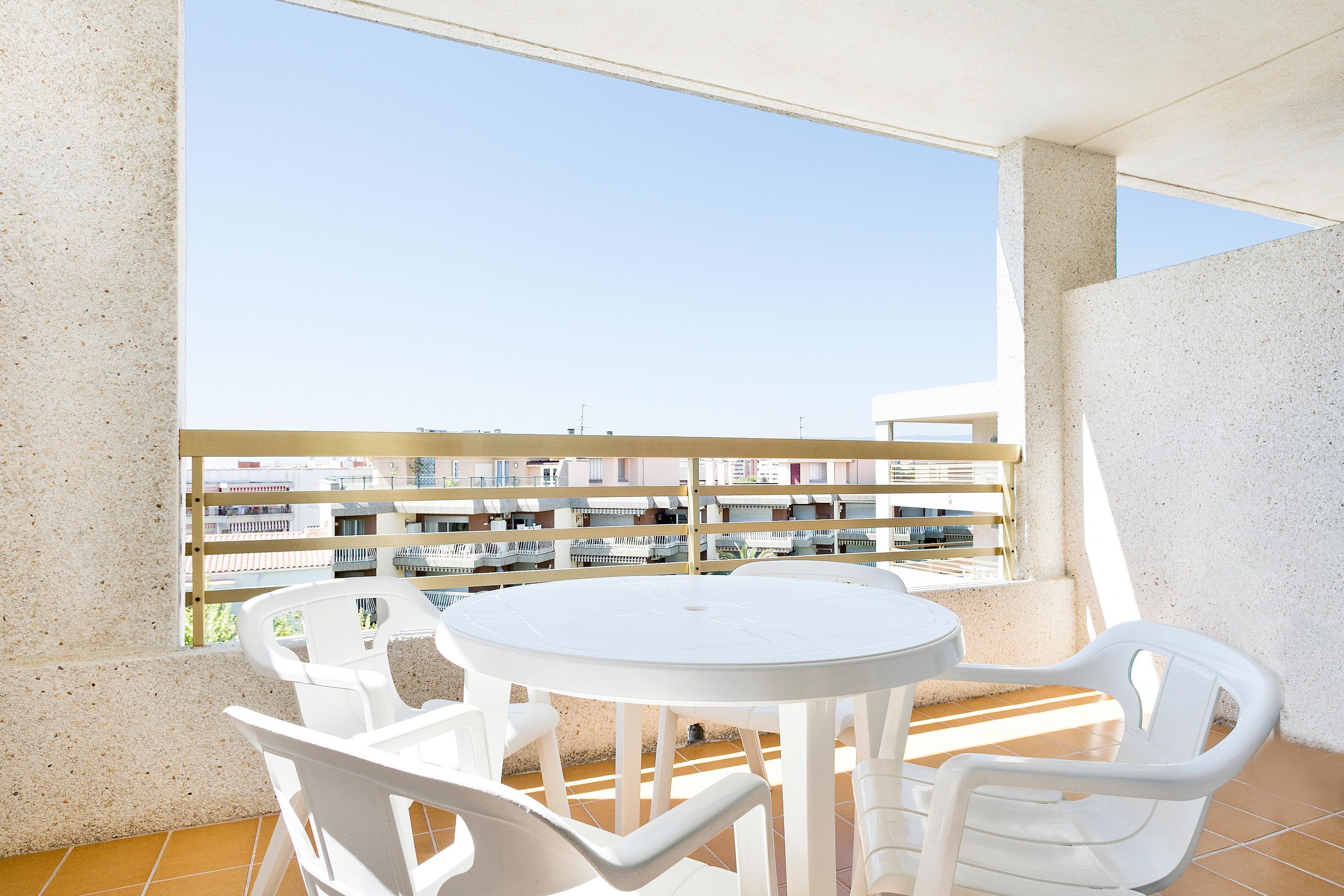 Een balkon met een witte tafel en stoelen, een uitzicht op gebouwen in de verte, en een hekwerk.