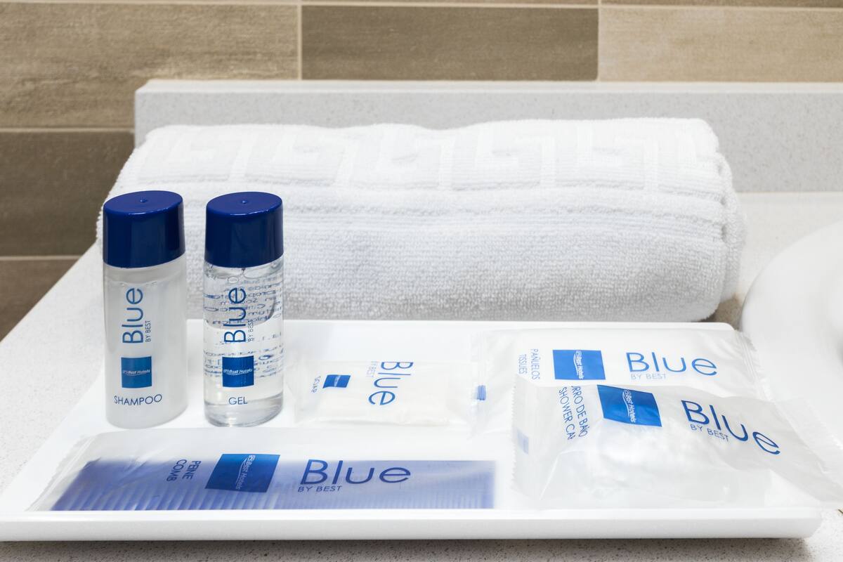 A bathroom with a white towel, two bottles of Blue shampoo and gel, and a tray with Blue shower gel and a bar of soap.