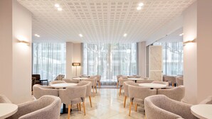 Un espace de restauration moderne avec des tables rondes et des chaises à revêtement gris, de grandes fenêtres avec des rideaux légers et un plafond orné d’un motif en réseau.