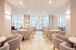 A modern dining area with round tables and grey upholstered chairs, large windows with sheer curtains, and a ceiling with a grid pattern.