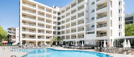 Un hotel de varios pisos con piscina, mobiliario exterior y un cielo azul despejado.
