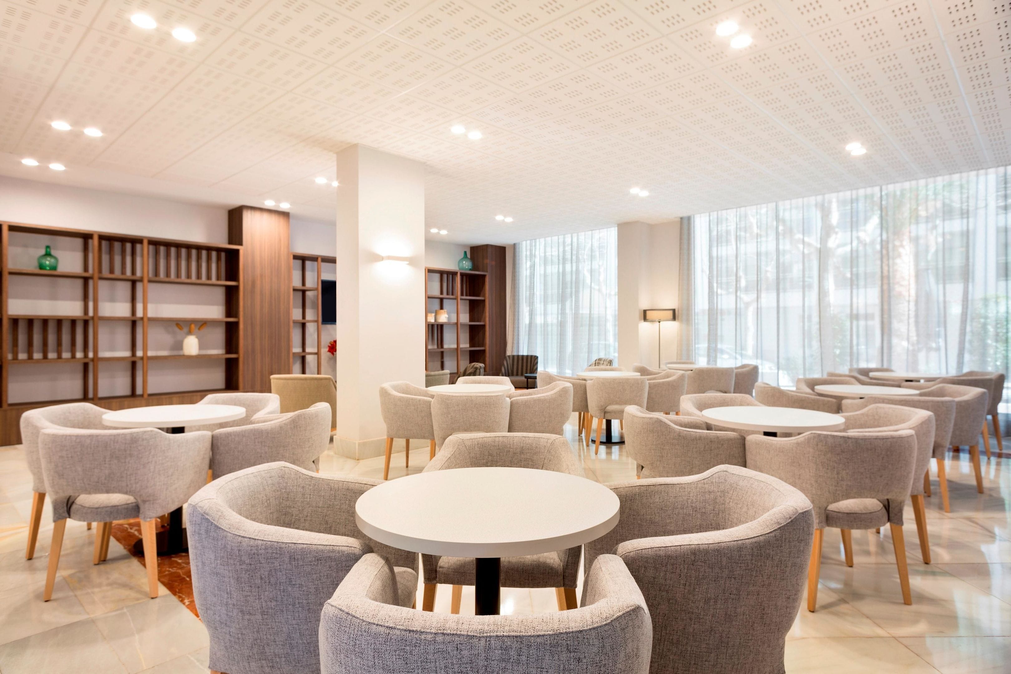 A modern interior with round tables and grey upholstered chairs, wooden bookshelves, and large windows allowing natural light.