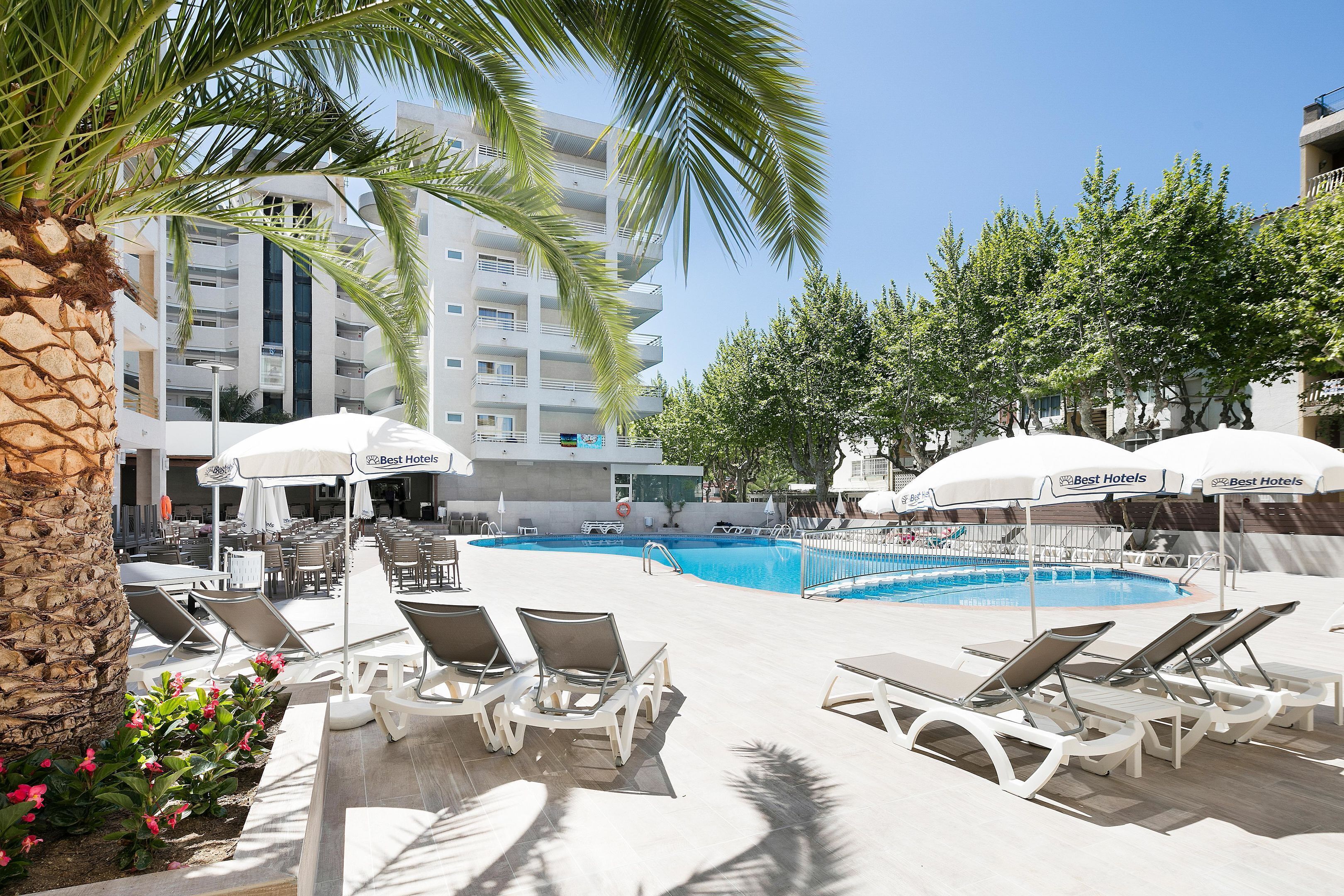 A pool area with lounge chairs and umbrellas, a clear blue pool, and a building with balconies in the background.