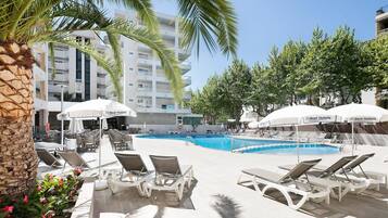 A pool area with lounge chairs and umbrellas, a clear blue pool, and a building with balconies in the background.