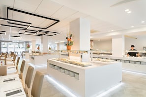 A modern dining area with white tables and chairs, a central buffet station, and a large potted plant.