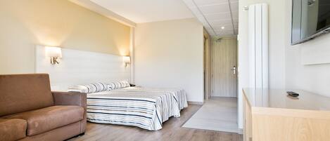 A modern hotel room with a sofa, a bed with striped bedding, a wooden desk, and a flat-screen TV mounted on the wall.