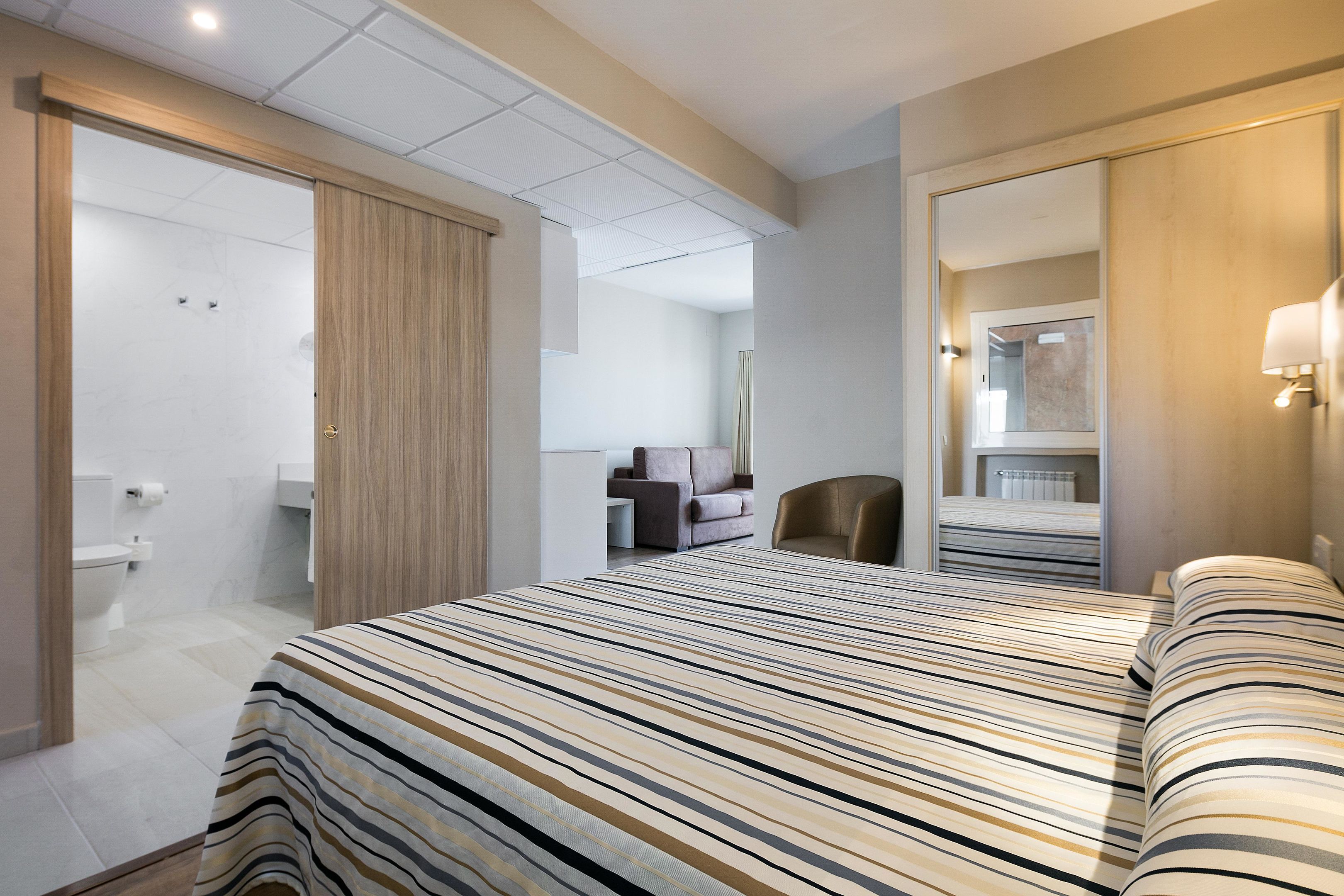 Una camera d'hotel con un letto grande, un angolo salotto con divano e poltrone, un bagno con il water e un lavandino, e uno specchio.