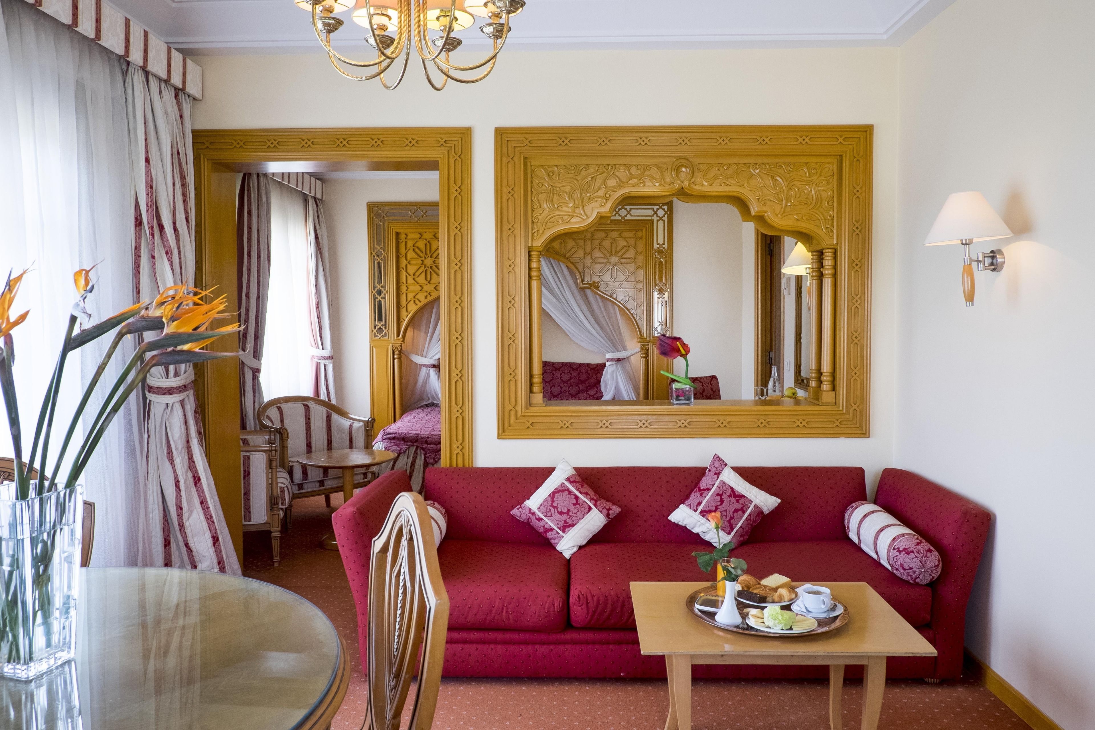 Una habitació amb un sofà vermell, una taula de vidre amb un plat de menjar, un mirall amb una estructura ornamentada i un penjallum.