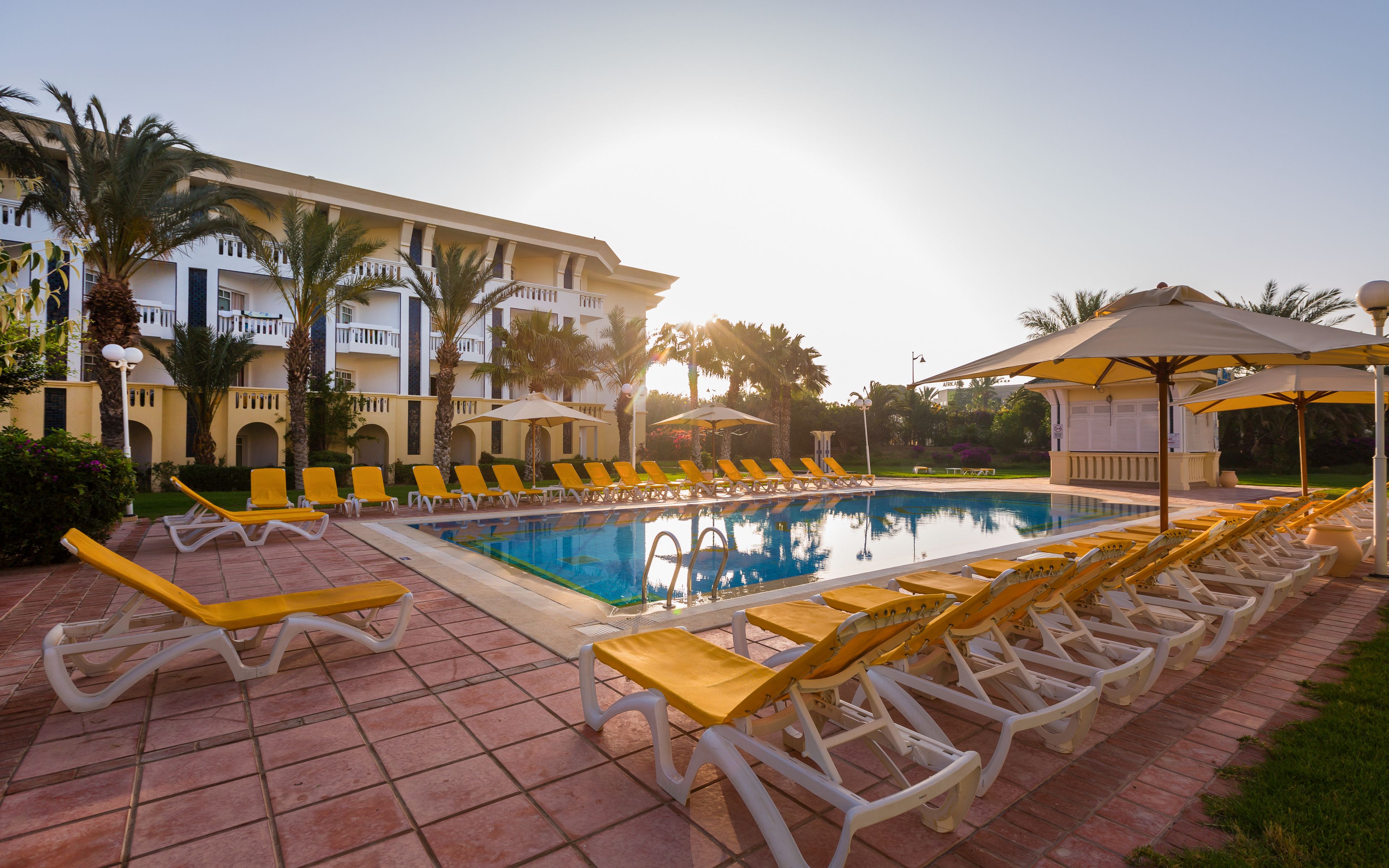 A poolside area with sun loungers, umbrellas, and a building with balconies.
