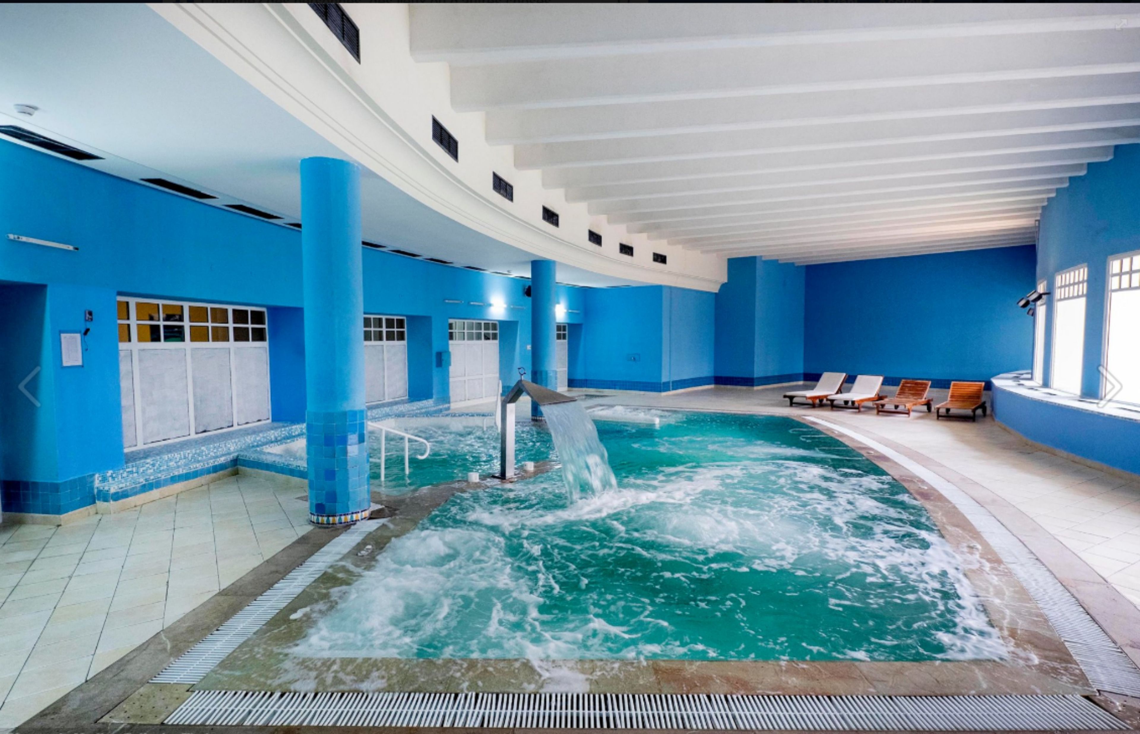 Une piscine intérieure aux murs bleus, agrémentée d’une cascade, et entourée de chaises longues.