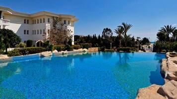 A large outdoor swimming pool with clear blue water, surrounded by rocks and palm trees, with a building in the background.