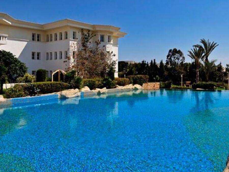 A large outdoor swimming pool with clear blue water, surrounded by rocks and palm trees, with a building in the background.