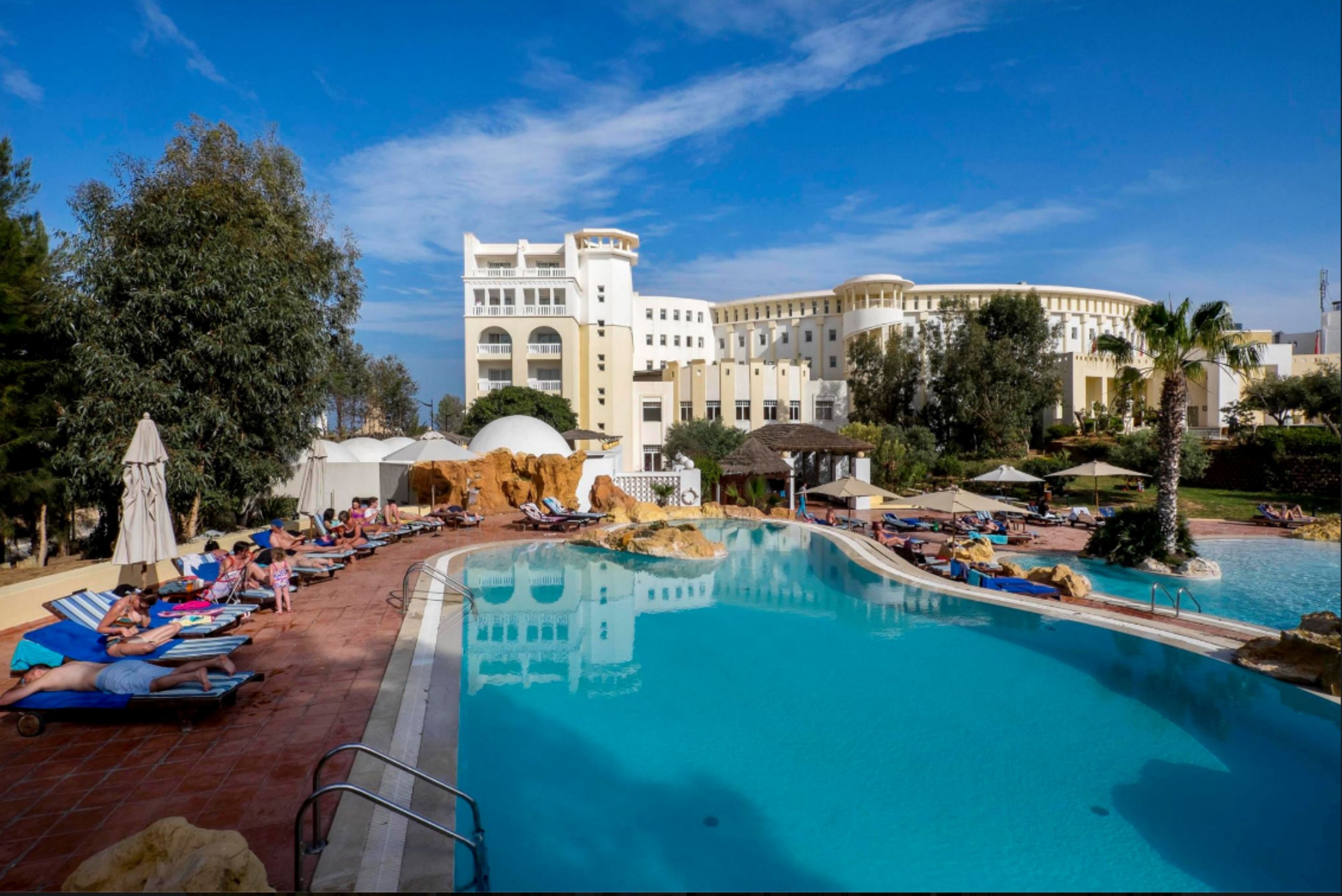 A resort pool area with sun loungers and umbrellas, surrounded by a large building with multiple balconies and a clear blue sky.