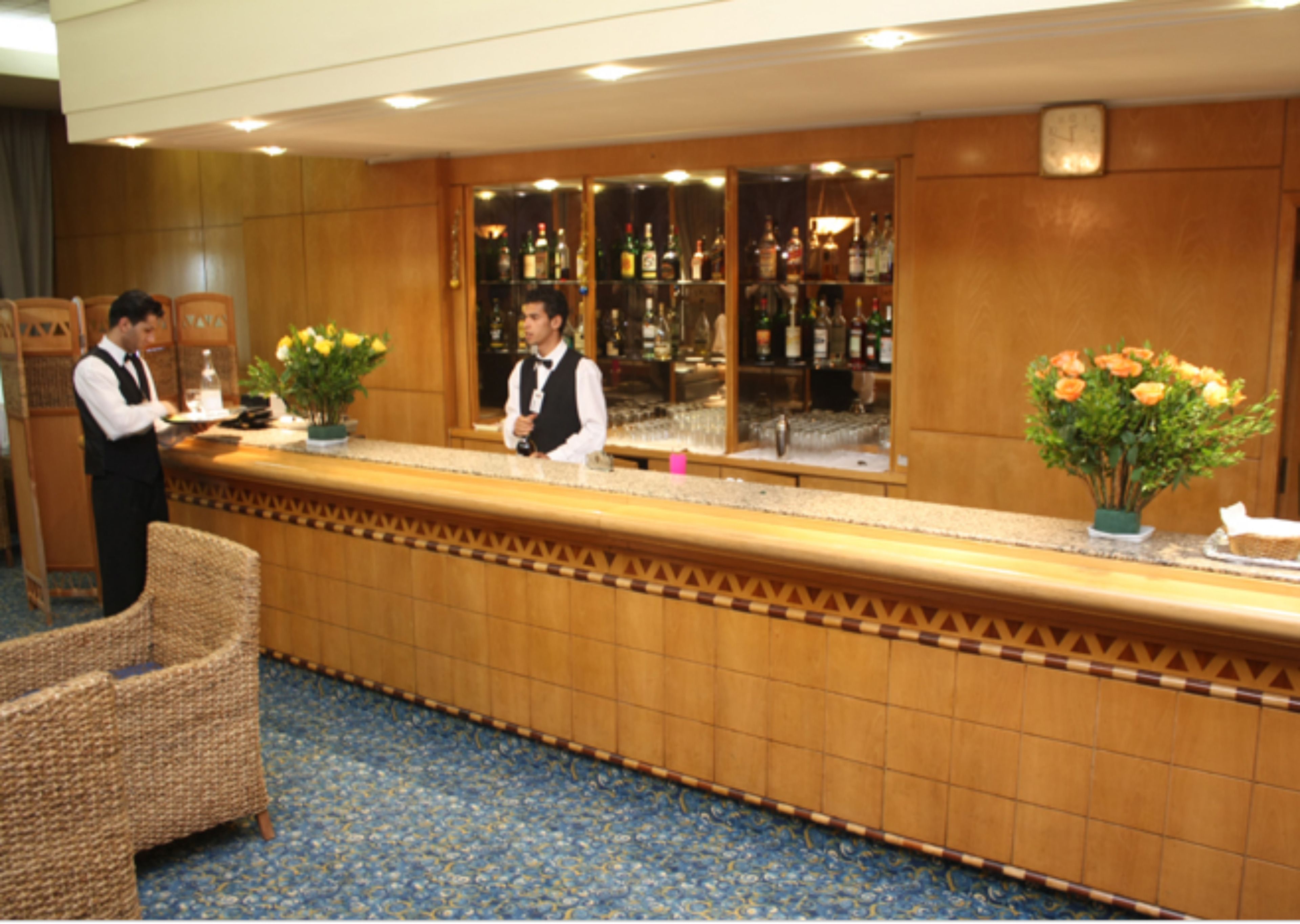 A hotel reception area with a counter, two waiters, a bar with bottles, and a clock on the wall.