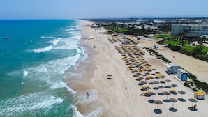 An aerial view of a beach with a row of beach umbrellas and sun loungers, a small boat on the water, and buildings in the background. - Medina Belisaire &Thalasso (Hammamet)