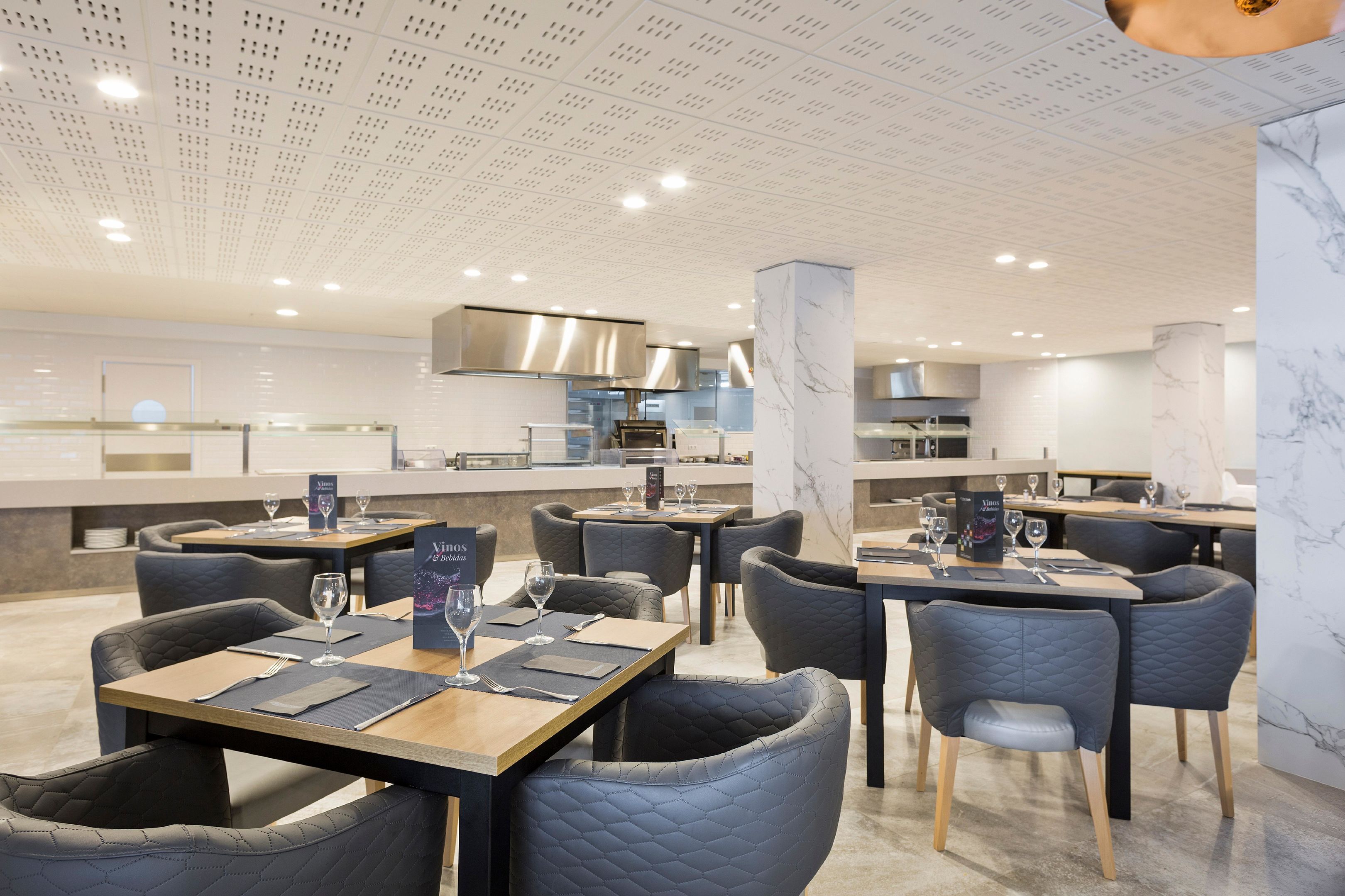 Un restaurante moderno con mesas y sillas, una cocina al fondo y un techo con un diseño único.