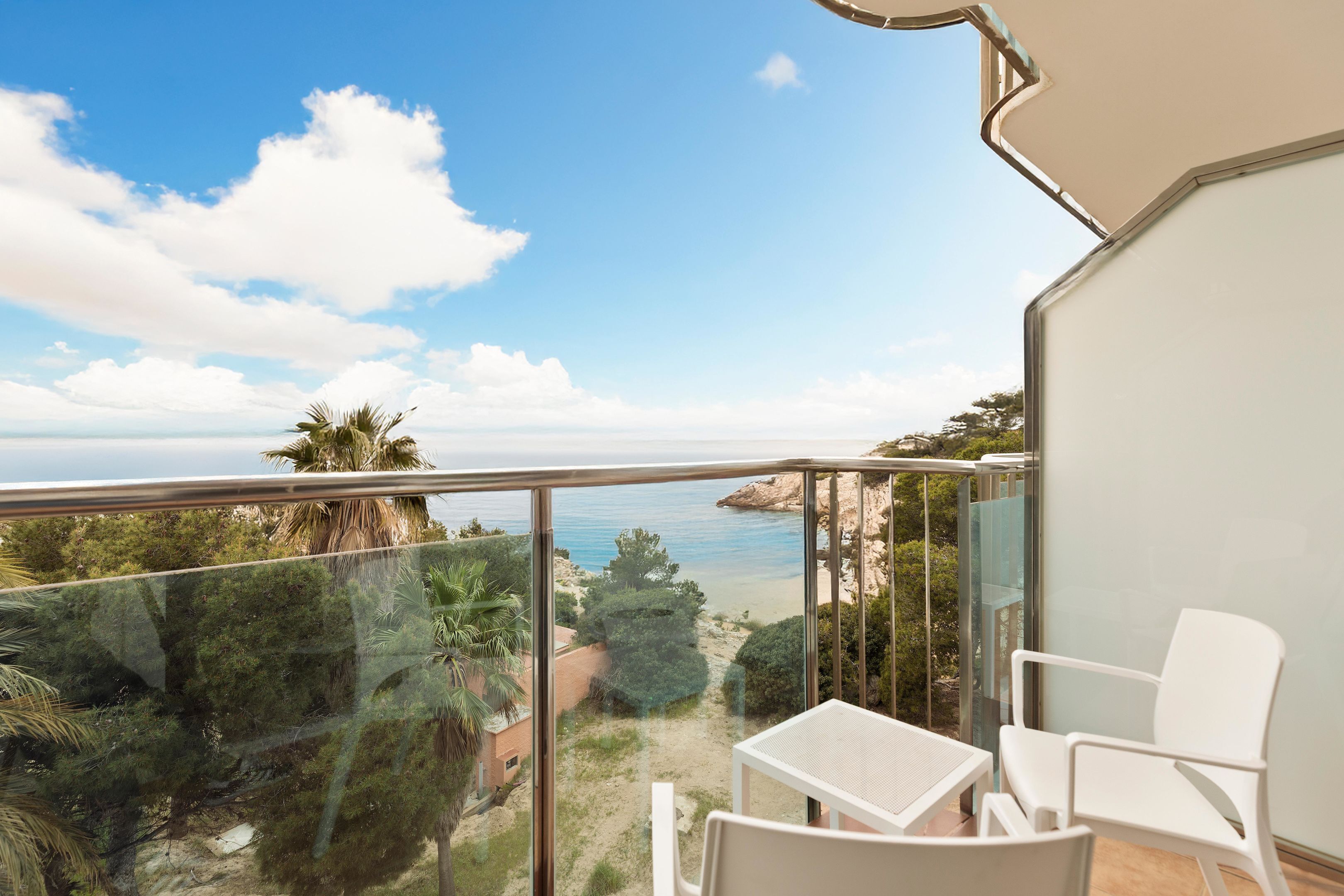Un balcón con barandilla de vidrio, sillas blancas y vistas al mar y la vegetación.
