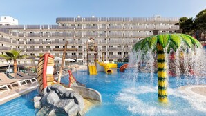 Una piscina con elementos acuáticos, una palmera y un edificio con balcones al fondo.