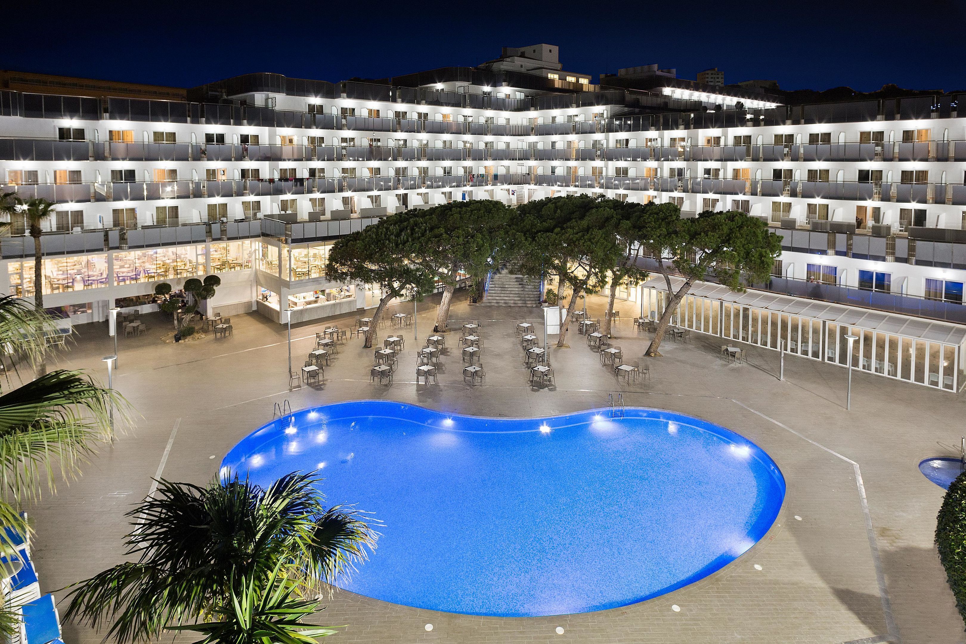 Un hotel con varios pisos, piscina, mobiliario exterior y una zona cubierta con mesas y sillas.