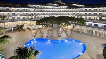 Un hotel con varios pisos, piscina, mobiliario exterior y una zona cubierta con mesas y sillas.