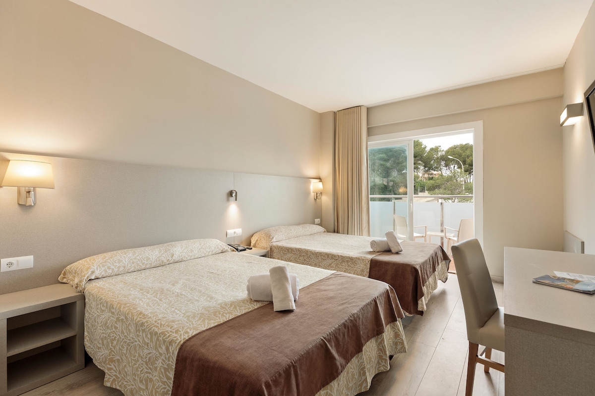 A hotel room with two beds, a desk, and a window with a view of trees.