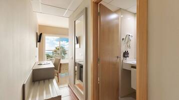 A hallway leading to a bathroom with a sink, a mirror, and a shower. There is a desk with a chair and a window with a view of palm trees.