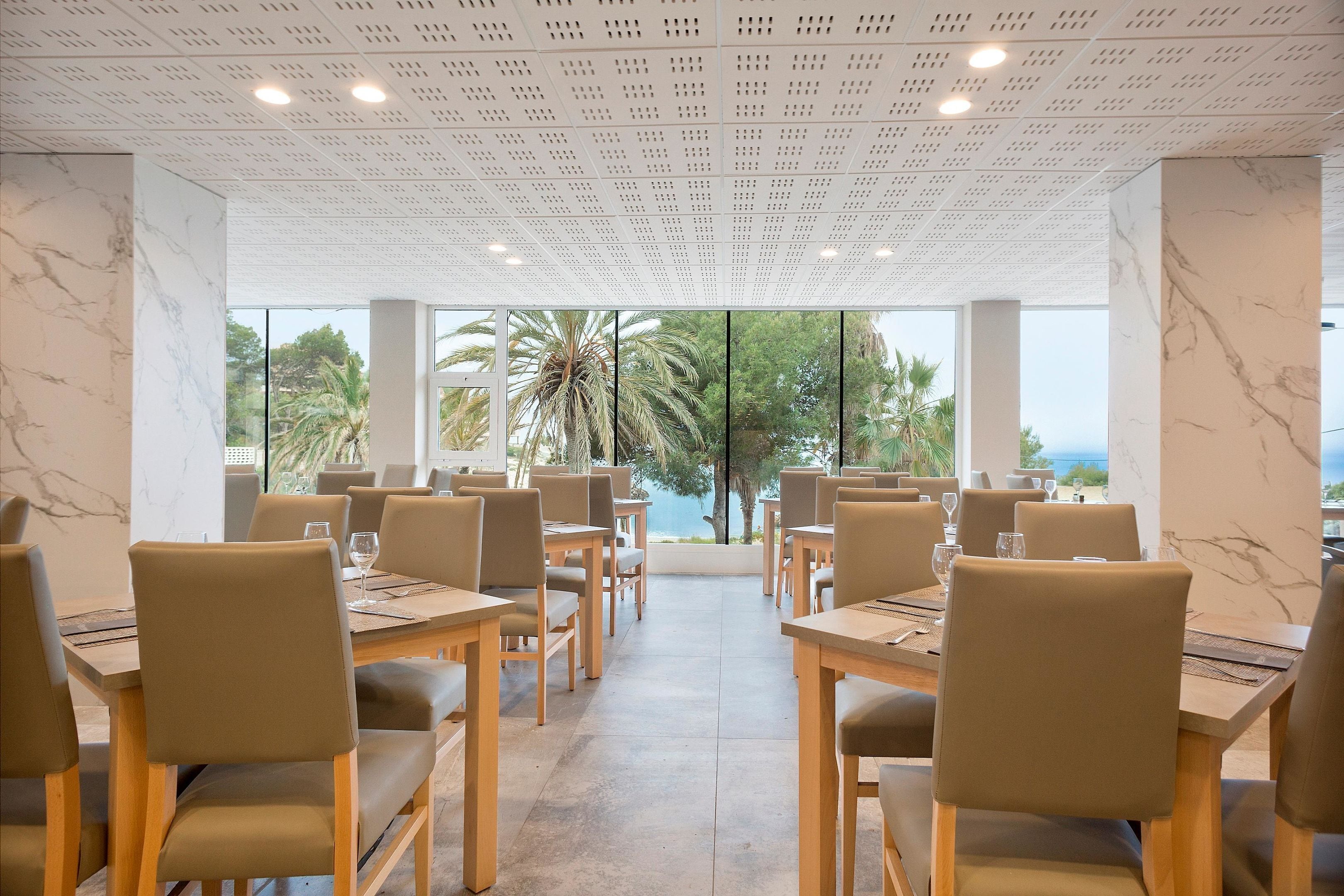 Un restaurante con paredes de mármol, mesas y sillas de madera, y vistas a palmeras y al mar.
