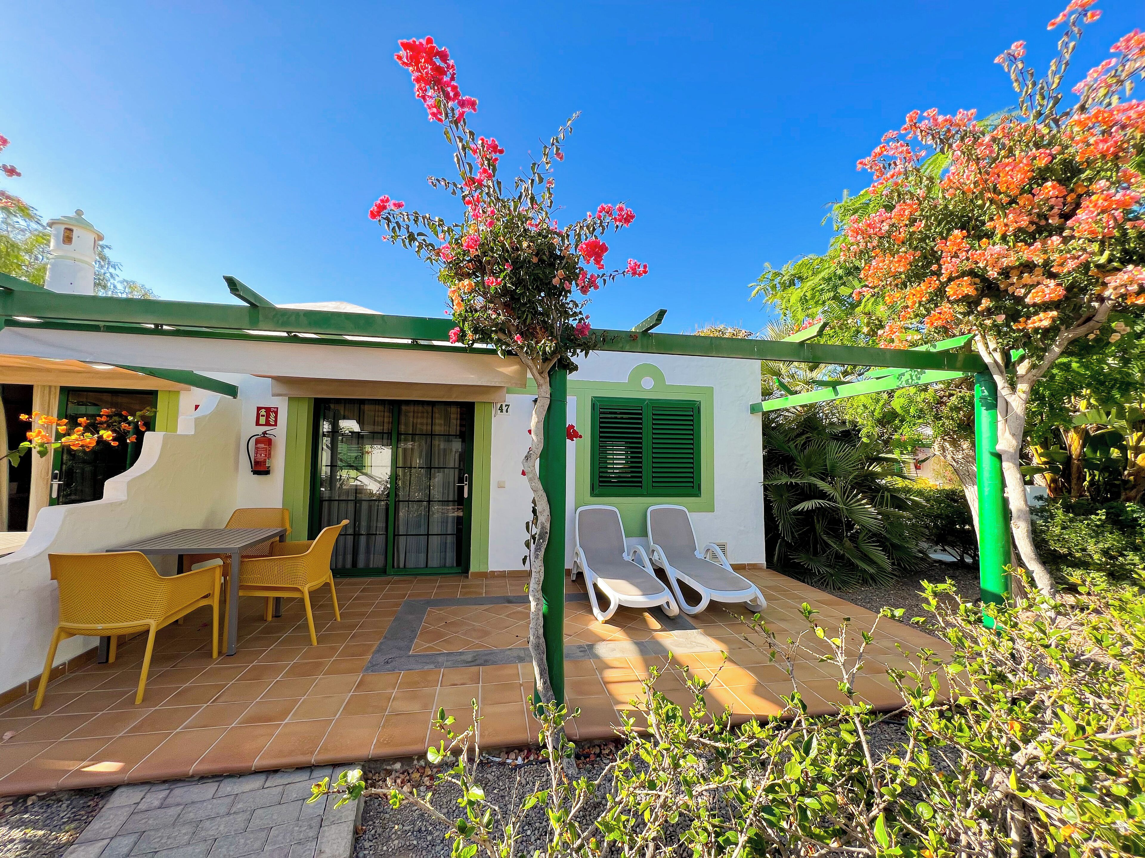 Un patio con mobiliario exterior, una pérgola verde y un edificio blanco con puerta verde.