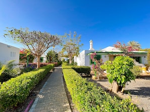 Un chemin mène à un bâtiment à la façade blanche et aux touches de vert, entouré de jardins et d’arbres bien entretenus.