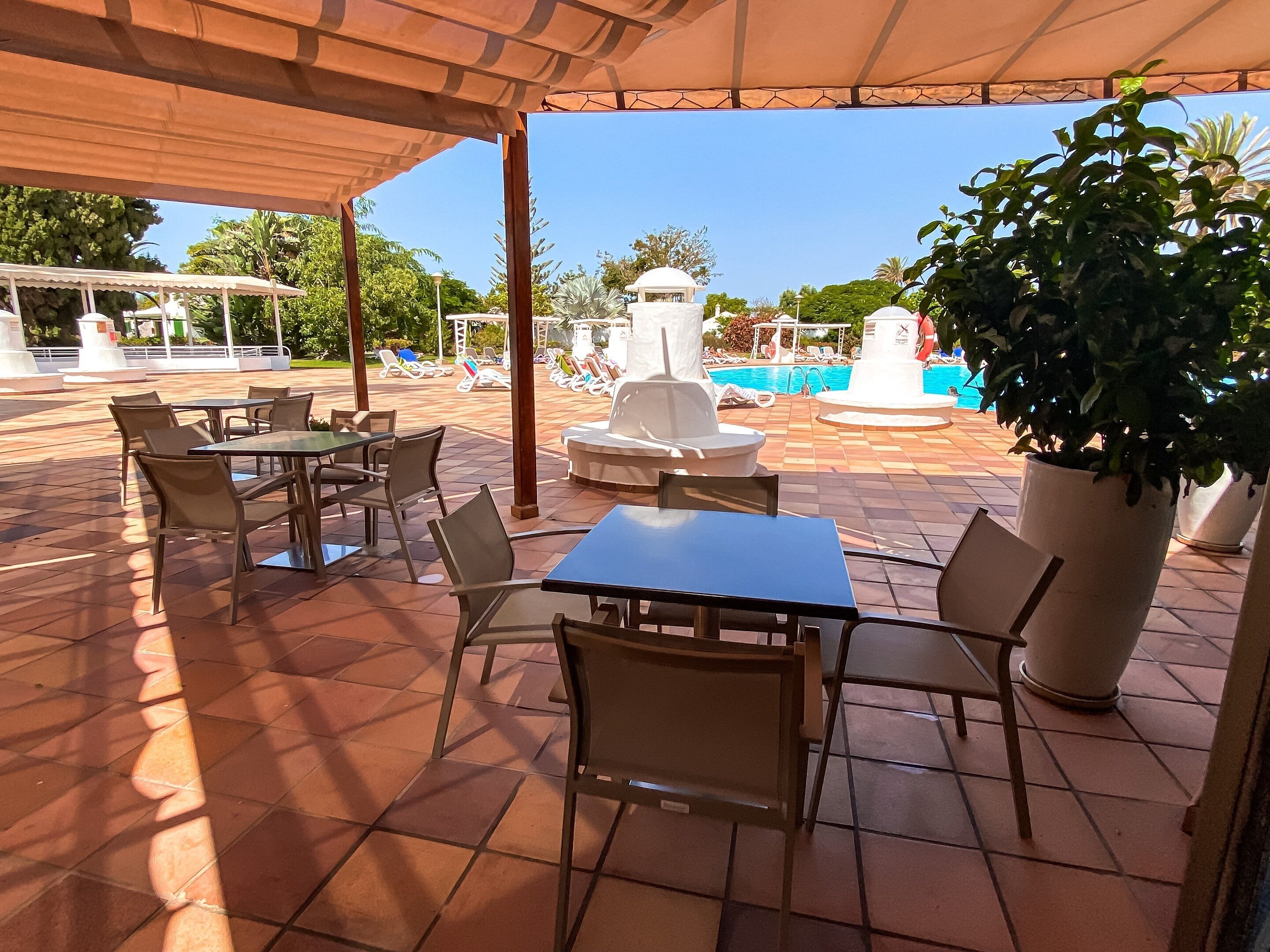 An outdoor patio area with tiled flooring, tables, and chairs. There is a pool with lounge chairs and a fountain in the background.