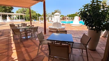 An outdoor patio area with tiled flooring, tables, and chairs. There is a pool with lounge chairs and a fountain in the background.