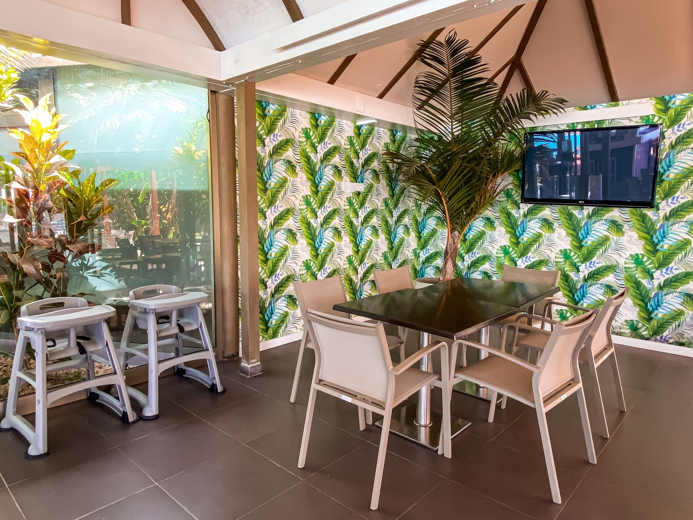 A modern dining area with a glass partition, a tropical leaf-patterned wall, a large flat-screen TV, and a central table with chairs.