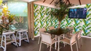 A modern dining area with a glass partition, a tropical leaf-patterned wall, a large flat-screen TV, and a central table with chairs.