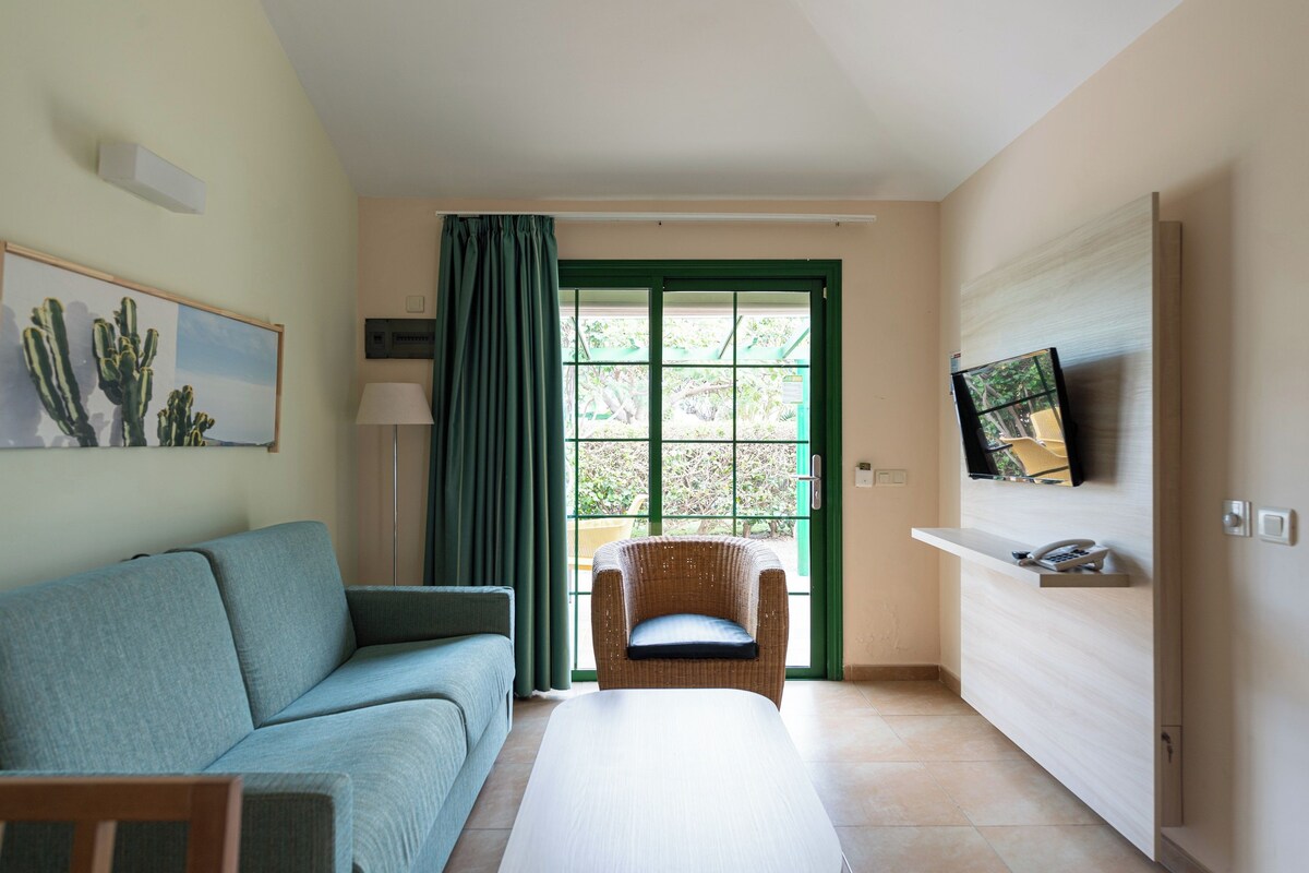 A living room with a sofa, a chair, a TV mounted on the wall, and a window with curtains.