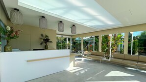 A modern reception area with a white reception desk, a seating area with benches, and a glass partition. - Bungalows Cordial Sandy Golf (San Bartolomé de Tirajana)