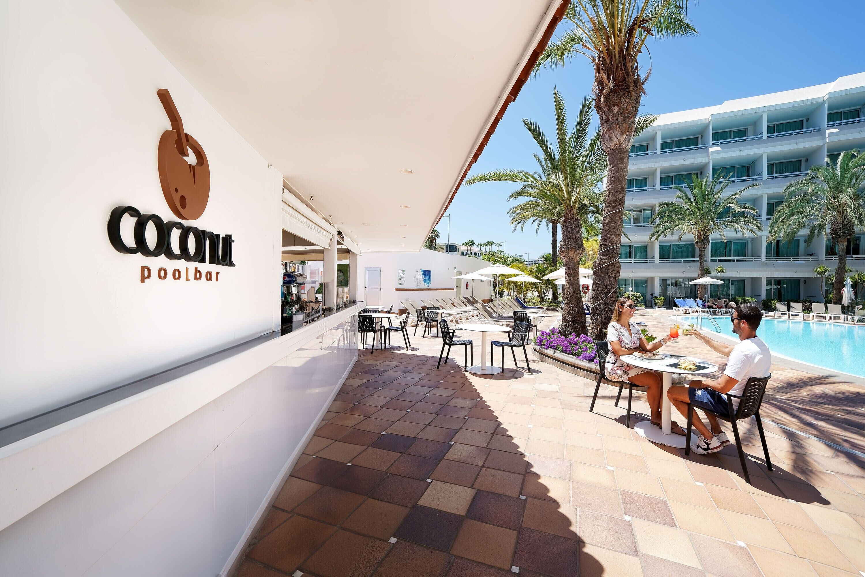 An outdoor pool area with a 'Coconut Poolbar' sign, palm trees, and a clear blue sky.